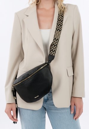 Persoon die een beige blazer en lichtblauwe jeans draagt met een zwarte, getextureerde crossbodytas met een zwart-beige patroon op de hengsel.