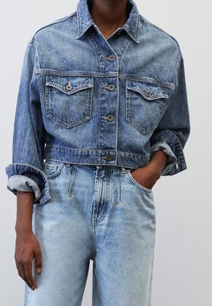 Personne portant une veste en jean bleue avec les manches retroussées et un jean taille haute bleu clair, debout avec une main dans la poche.