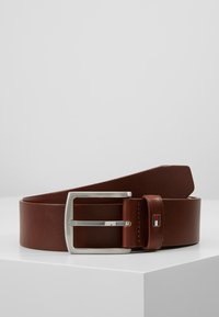 Ceinture en cuir brun avec une boucle rectangulaire argentée, présentant un petit logo en rouge, blanc et bleu sur le passant. Texture lisse.