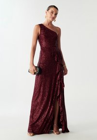 Jedno rameno burgundské večerní šaty s flitry, s kaskádovou volánovou sukní, vysokým rozparkem a vypasovaným pasem. Doplněné černým psaníčkem.