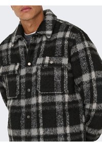 Homme portant une veste en laine à carreaux noirs et gris avec deux poches à rabat et des boutons-pression en métal, visible du cou jusqu'au milieu du torse.