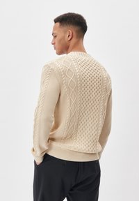Beige kötött pulóver, amely texturált mintákkal, fonott designnal, bordázott mandzsettákkal és kerek nyakú kivágással rendelkezik, laza szabású és közepes vastagságú.