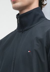 Άνδρας που φοράει ένα μαύρο τζάκετ με φερμουάρ και ψηλό κολάρο, με ένα μικρό κόκκινο, λευκό και μπλε λογότυπο στο στήθος, κοιτάζοντας δεξιά.