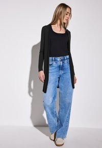 Svart långärmad cardigan över en åtsittande svart topp, i kombination med ljusblå vida jeans och beige sneakers. Släta texturer och avslappnad stil.
