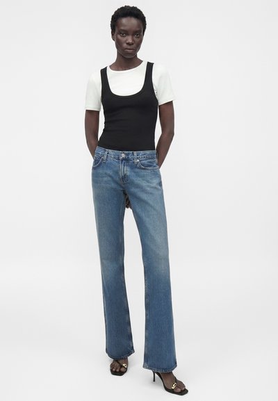 Γυναίκα που φοράει λευκό κοντομάνικο τοπ κάτω από μαύρο αμάνικο, μπλε ψηλόμεσο τζιν κανονικής γραμμής και μαύρα πέδιλα με τακούνι και χρυσές λεπτομέρειες.