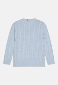 Lys blå kabelstrikket genser med ribbestrikkede mansjetter og nederkant, med en V-formet halslinje og en liten rød, hvit og blå detalj på baksiden.