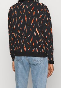 Černá mikina se vzorem oranžových a červených blesků, s žebrovanými manžetami a lemem, spojená s světle modrými džíny.