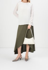 Haut blanc à manches longues avec manches texturées, jupe longue asymétrique vert olive, escarpins gris à bout pointu, et un sac à main blanc avec des détails en dentelle.
