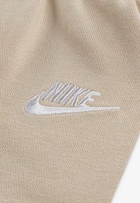 Bežinė medžiaga su tekstūruotu audiniu turi siuvinėtą sidabrinį Nike logotipą, kuriame išryškėja išskirtinis swoosh ir ryškus užrašas.