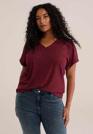 Frau mit lockigem dunklem Haar, die ein strukturiertes bordeauxfarbenes V-Ausschnitt-Kurzarmshirt und blaue Jeans trägt, steht vor einem schlichten Hintergrund.