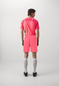 Maglietta da sport rosa brillante con motivo geometrico, pantaloni corti abbinati, calze bianche e scarpette nere. Vista posteriore.
