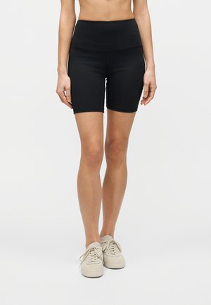 Nainen, joka pukeutuu korkeavyötäröisiin mustiin pyöräilyshortseihin ja beigeihin lenkkareihin, seisoo yksinkertaisen vaalean taustan edessä.