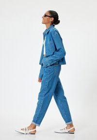 Conjunto de chaqueta de mezclilla azul y pantalones con un corte cropped y puños enrollados. El modelo lleva puestas zapatillas blancas con detalles de hebilla y gafas de sol oscuras.