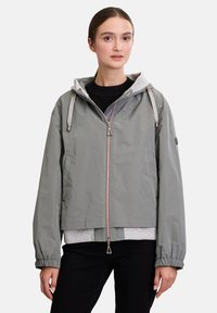 Lichtgrijze jas van waterafstotend materiaal met een capuchon, elastische manchetten en een rits aan de voorkant. Bevat een contrasterende grijze binnenvoering.