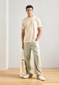 Beige cotton t-shirt, relaxed fit, paired with light blue loose-fit jeans. The person holds a beige tote bag and wears white sneakers.