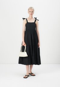 Melnā slāņveida vasaras kleita ar plānām siksnām un loku akcentiem, saskaņota ar mazu baltu rokassomu un melniem flip-flops. Gluda auduma tekstūra.