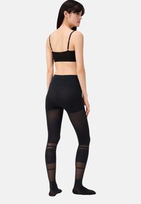 Schwarze Leggings mit durchsichtigen Streifen, hohem Taillenbund und leichtem Material. Das Model trägt einen schlichten schwarzen Bralette, hat die Haare offen und steht seitlich.