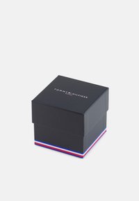 Boîte en carton noir avec un couvercle arborant le logo blanc "TOMMY HILFIGER". La base présente un accent rayé rouge, blanc et bleu tout autour.