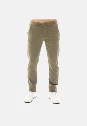 Pantaloni verde oliva aderenti indossati da una persona con scarpe da ginnastica bianche, mostrati su uno sfondo chiaro e uniforme.