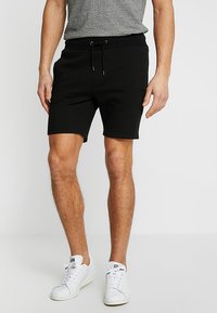 Schwarze Baumwollshorts mit elastischem Bund und Kordelzug. Sie verfügen über Seitentaschen und eine leicht zulaufende Passform, kombiniert mit weißen Sneakern.