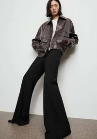 Brązowa skórzana kurtka z dwiema przednimi kieszeniami, zamek błyskawiczny, i ściągacz na dole; zestawiona z czarnymi spodniami o szerokich nogawkach. Gładka struktura na całej powierzchni.