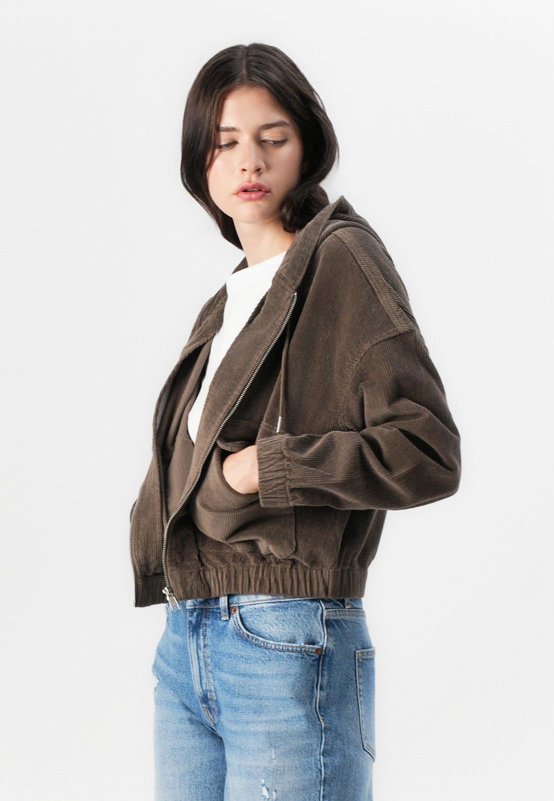 Veste bomber en velours côtelé marron avec capuche, zip à l'avant et poches latérales, associée à un jean bleu clair. Coupe structurée avec taille élastique.
