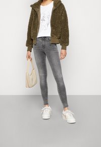 Osoba ubrana w oliwkową, puszystą kurtkę na zamek, białą koszulkę z grafiką, szare jeansy skinny i białe trampki, trzyma małą beżową torebkę.
