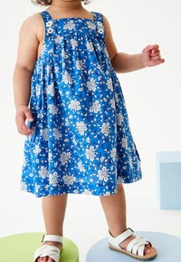 Sininen kukkakuosiset mekko, jossa on valkoisia kukkia, neliökaula-aukko, nappikoristeet olkaimissa ja rypytetty vyötärö. Käytetty valkoisten sandaalien kanssa värikkäillä palkeilla.