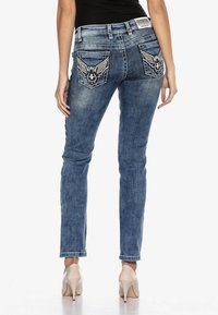 Schmale Jeans aus mittlerem blauen Denim mit einem verwaschenen Finish. Flügelstickerei auf den hinteren Taschen. Modell kombiniert mit hautfarbenen High Heels.