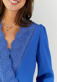 Helle blaue Bluse mit einem gewellten Spitzenkragen, der mit Stickdetails verziert ist, und puffartigen Ärmeln. Glatter Stoff mit feinen Spitzelementen.