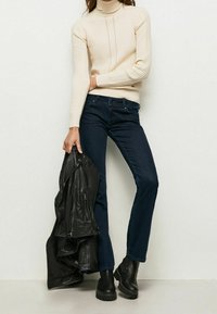 Pull en laine crème avec col roulé côtelé, jeans évasés bleu foncé, veste en cuir noire portée sur le bras et bottines noires chunky.