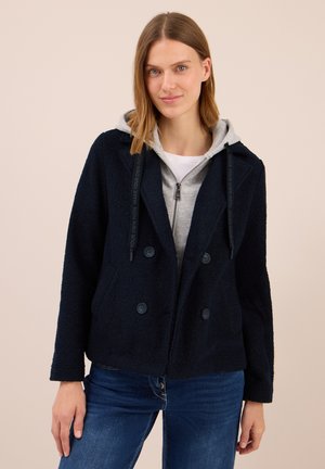 Femme portant une veste bleue marine à boutons sur un sweat à capuche gris avec des cordons, associée à un jean bleu, se tenant devant un fond beige.