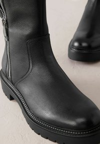 Bottines en cuir noir avec une finition texturée, fermeture éclair sur le côté et semelle robuste. Elles présentent un accent cousu autour de la base.