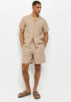 Młody mężczyzna z krótkimi włosami, ubrany w beżową, krótką koszulę na guziki, pasujące szorty i brązowe klapki, stoi z rękami w kieszeniach.