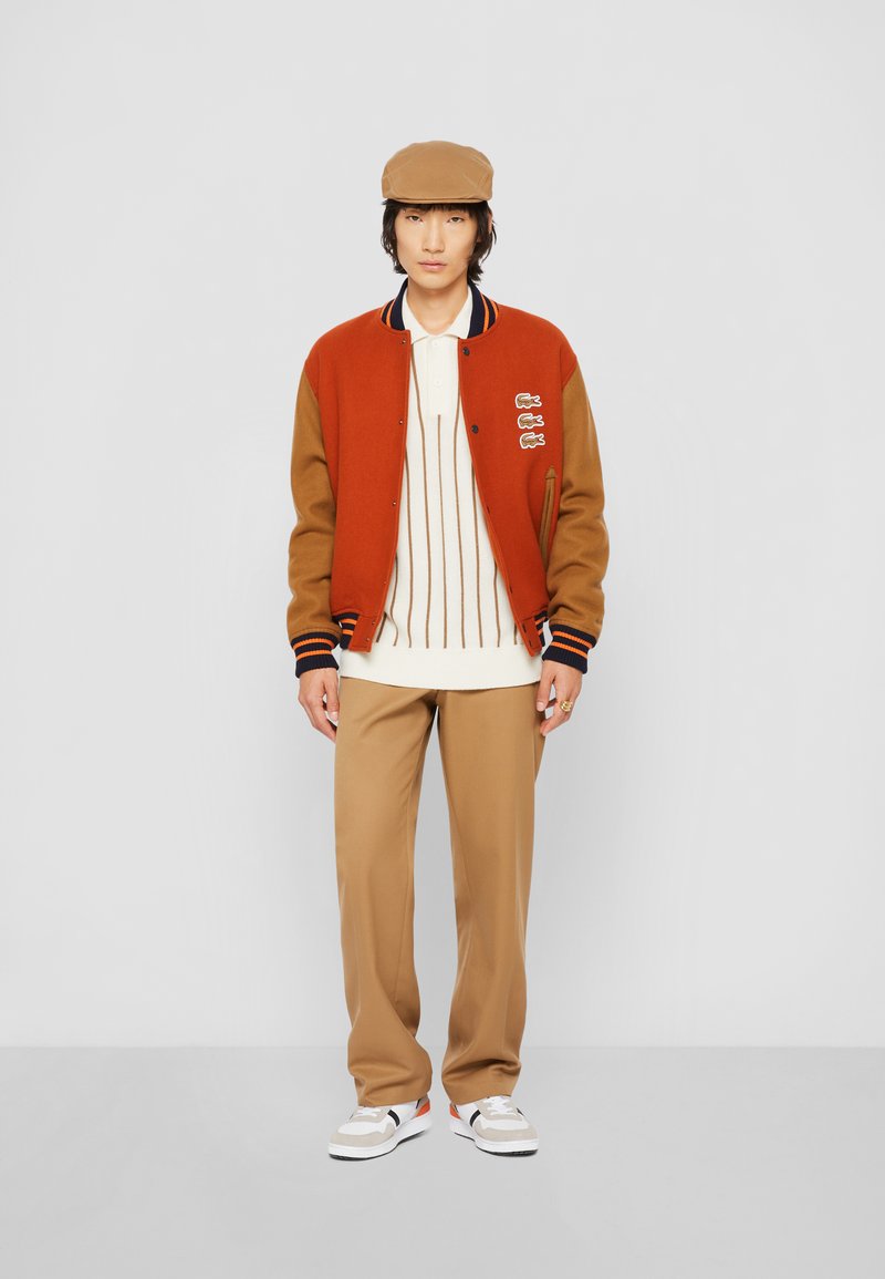 Chaqueta varsity marrón y naranja con rayas, camiseta polo crema, pantalones beige y zapatillas blancas con acentos en negro y rojo.