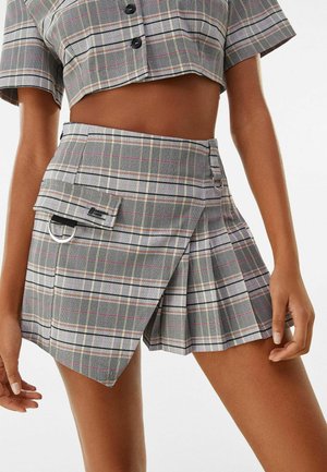 Femme portant un top court boutonné à carreaux gris et une mini-jupe plissée asymétrique assortie avec poche et anneaux métalliques.