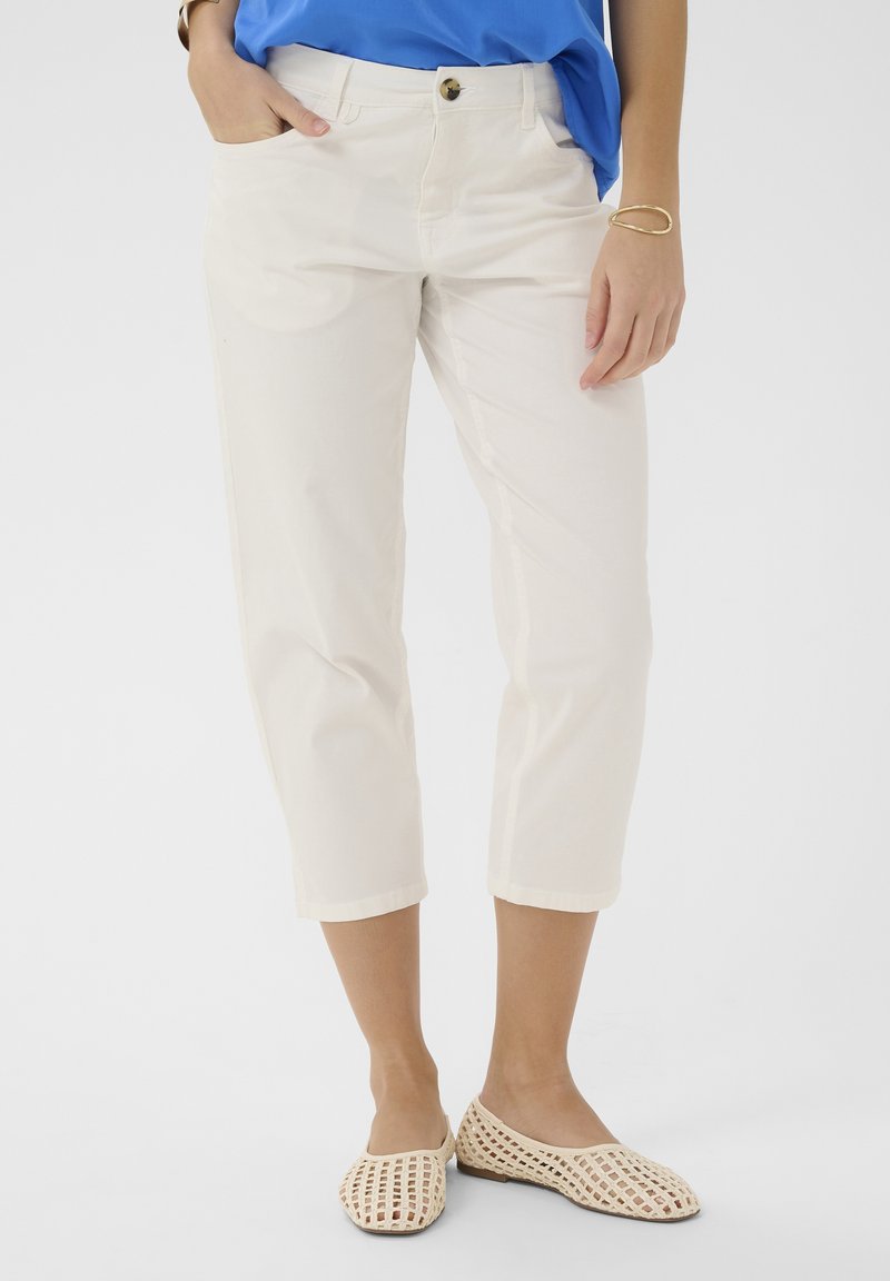 Mujer con pantalones blancos recortados, zapatos planos de tejido crema, blusa azul y pulsera dorada, de pie con una mano en el bolsillo.