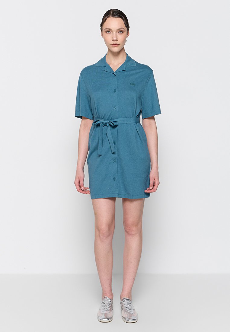 Turkusowa sukienka zapinana na guziki, z krótkimi rękawami, kołnierzykiem i paskiem w talii. Wykonana z miękkiego materiału o luźnym kroju. Dyskretne logo na klatce piersiowej.