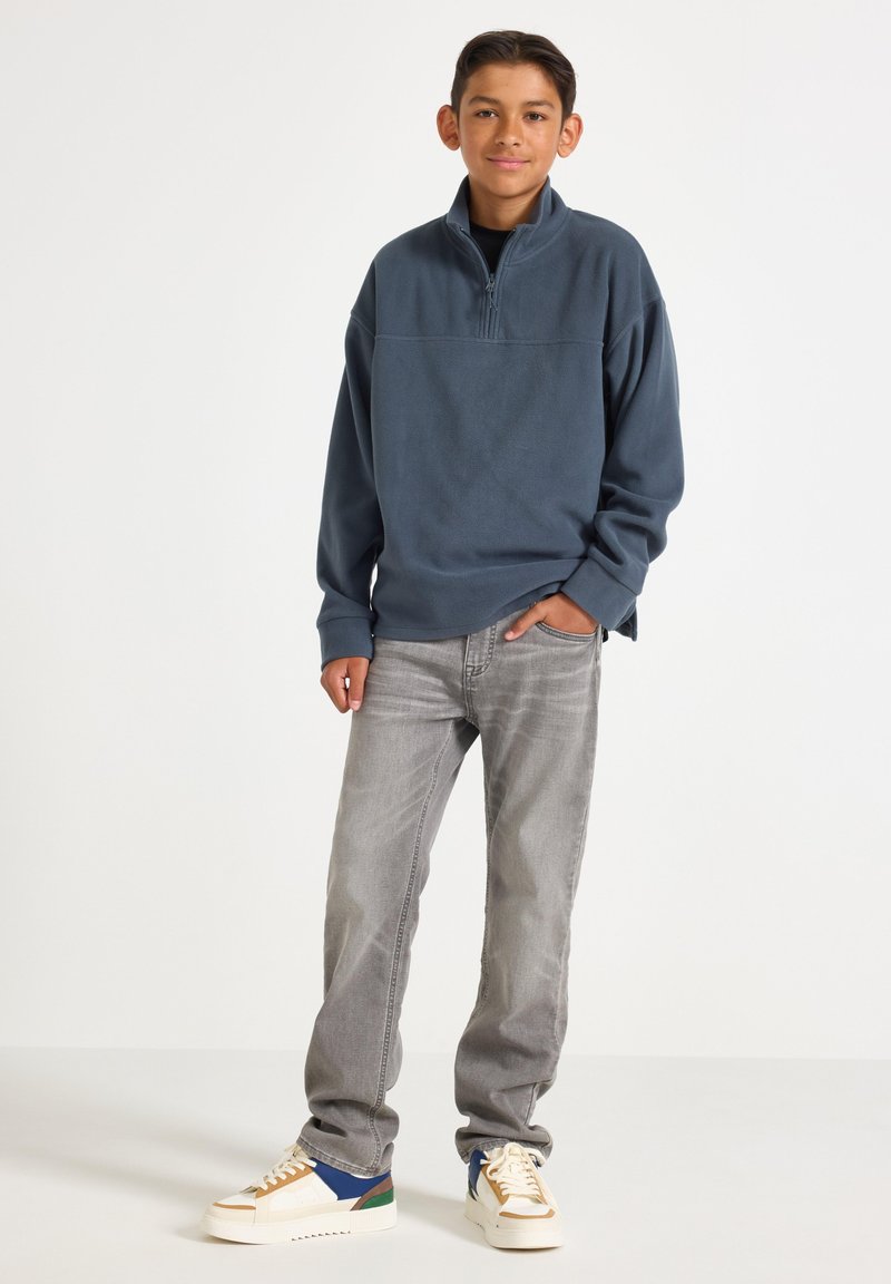 Fleecepullover med halvmatch i mörkblå, tillsammans med ljusgrå jeans och flerfärgade sneakers med blå, gröna och bruna detaljer.