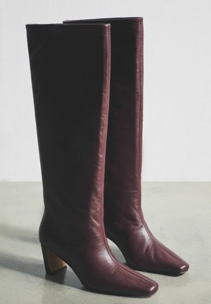 Burgundiapunased nahast põlveni saapad, millel on terav nina ja plokkkonts, sile tekstuur ning minimaalne õmblusdetailide kasutus.