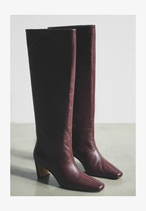 Burgundiapunased nahast põlveni saapad, millel on terav nina ja plokkkonts, sile tekstuur ning minimaalne õmblusdetailide kasutus.