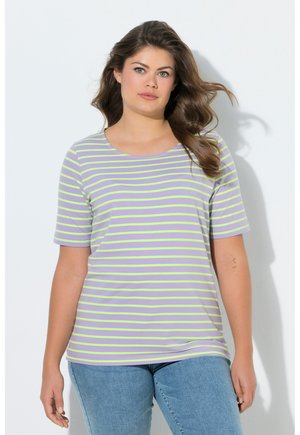 Frau mit langen braunen Haaren, die ein kurzärmeliges Hemd mit lavendelfarbenen und hellgrünen Streifen sowie eine blaue Jeans trägt, stehend vor einem weißen Hintergrund.