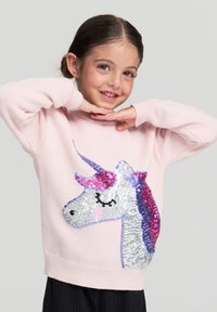Ragazzina sorridente con le mani sotto il mento, indossa un maglione rosa chiaro con un grande design di unicorno in paillettes e pantaloni neri.