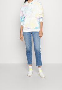 Hoodie tie-dye pastello con tasca a marsupio, cordini bianchi e polsini a costine, abbinato a jeans dritti blu chiaro e sneakers bianche.