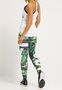 Legíny v živě zelené barvě s černým maskovacím vzorem a bílými akcenty. Uvolněný bílý top bez rukávů s detailní síťovinou. Černé sportovní boty.