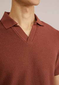 Rust-colored polo shirt with a textured diamond pattern, short sleeves, and a v-neck collar featuring a solid trim.