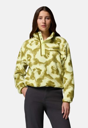 Kvinde iført en beige og olivengrøn mønstret fleecepullover med trykknapkrave og lynlåslomme på brystet, kombineret med mørke bukser.