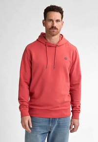 Korálová mikina s kapucí se stahovací šňůrkou, žebrovanými manžetami a lemem. Malé logo na hrudi. Pohodlná látka, volný střih, nošeno s světle modrými džíny.