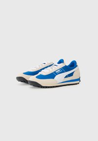 Scarpe atletiche blu e crema con tomaia in suede e tessuto, lacci bianchi e suole in gomma nera. Presentano un logo bianco prominente e dettagli.