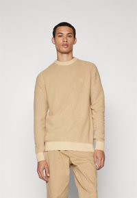 Beige gestrickter Pullover mit Rundhalsausschnitt, gerippten Bündchen und Saum. Besitzt ein subtil strukturiertes Muster und ein kleines Logo auf der Brust.